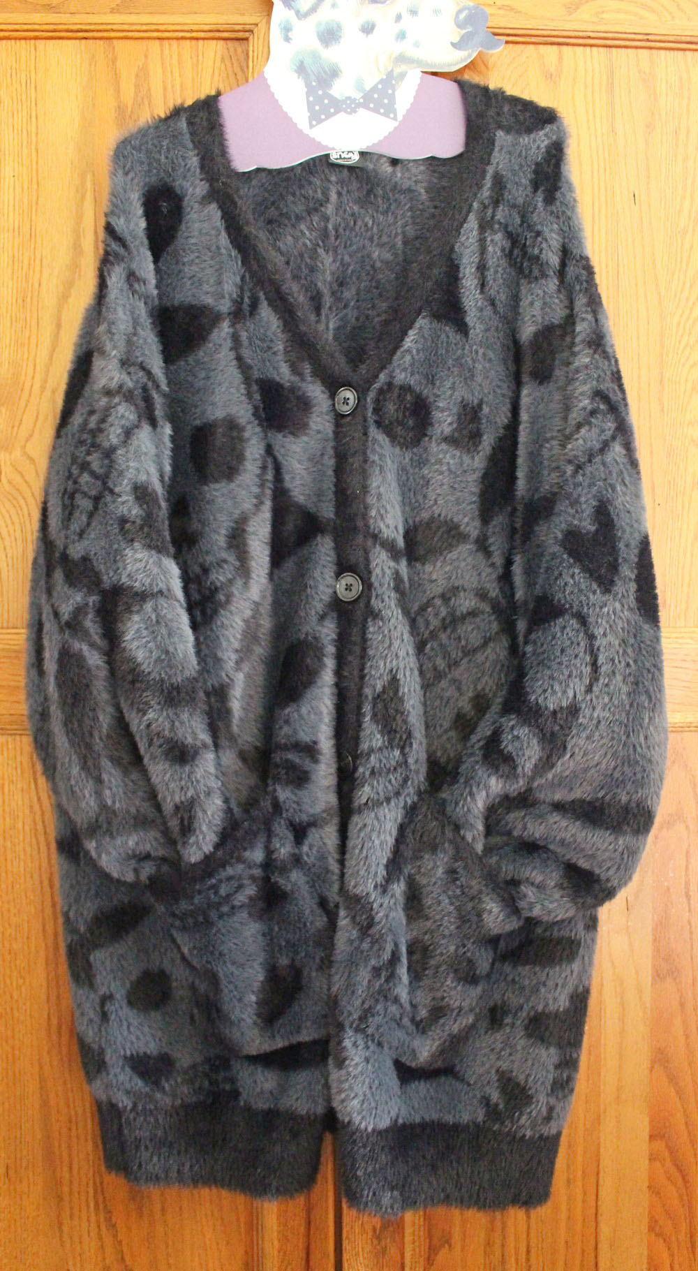 A fuzzy, oversized, dark grey cardigan with a black abstract pattern and black buttons, hanging on a wooden door—timeless and cosy, it’s the perfect example of Clothes That Last.