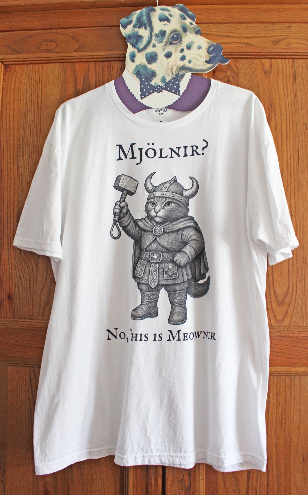 A white T-shirt hangs on a wooden door. The shirt features a cartoon cat dressed as a Viking, holding a hammer. Text above reads “Mjölnir?” and below reads “No, his is Meownir.”.