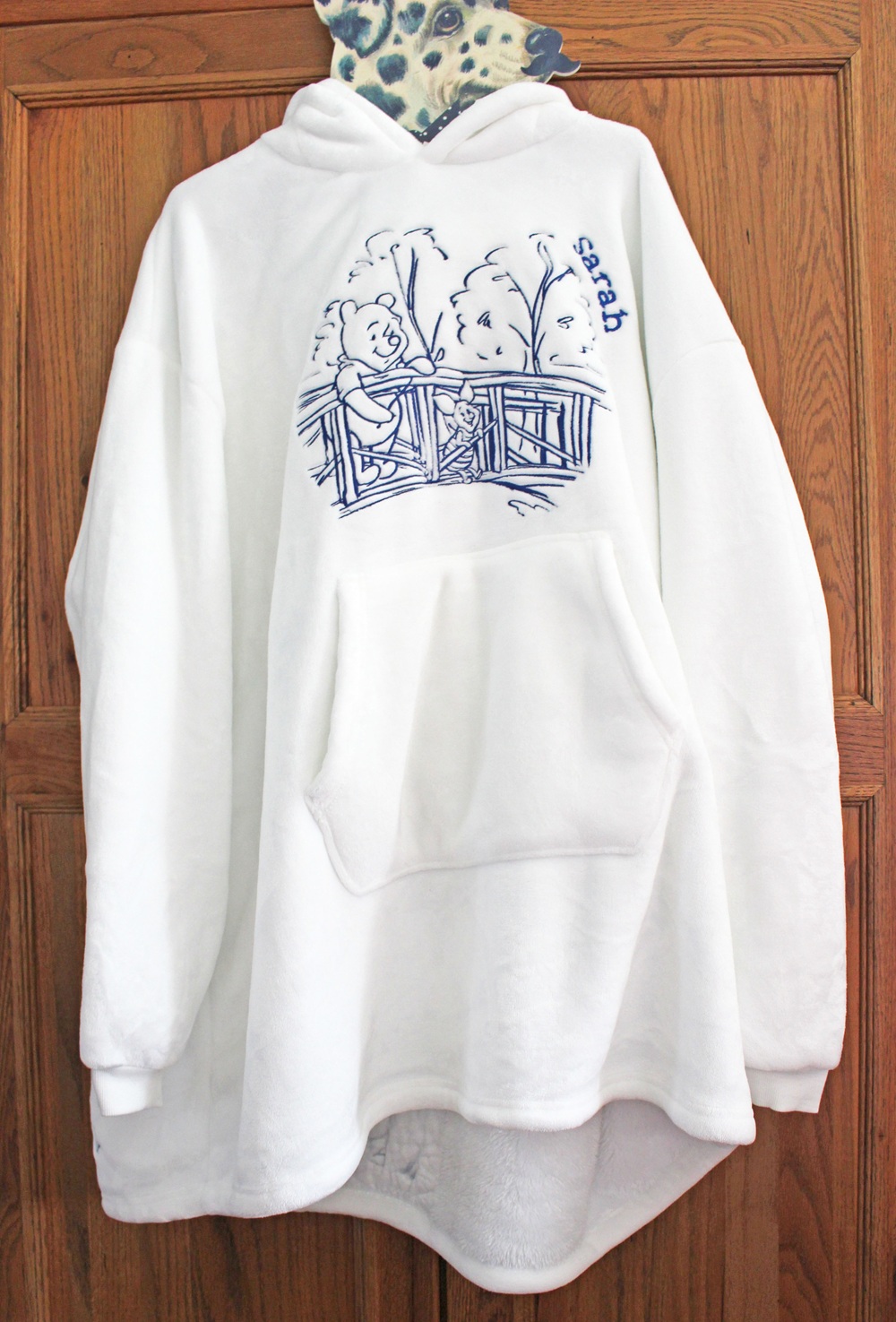 A white oversized hoodie with a front pocket, featuring a blue line drawing of Pooh Bear leaning on a wooden fence under trees, and the word “sad” written nearby, hanging on a wooden door.