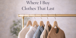 A gold clothes rail displaying neutral-coloured garments on wooden hangers with the text Where I Buy Clothes That Last above. The background is softly blurred with a hint of greenery.