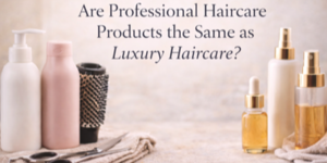 Several haircare products, including bottles, spray bottles, a dropper bottle, and a round hairbrush, are arranged on a light surface. Text above asks, Are Professional Haircare Products the Same as Luxury Haircare?.