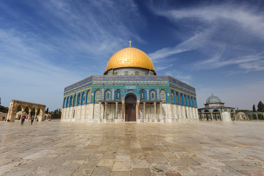 The Dome of the Rock, a historic Islamic shrine with a gold dome and ornate blue tilework, stands on a stone plaza under a partly cloudy sky in Jerusalem.