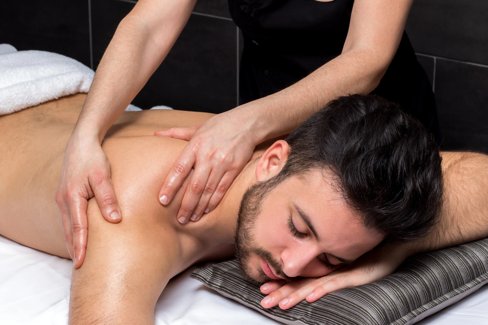 A man lies face down on a massage table with his eyes closed as a person massages his shoulder and upper back. He appears relaxed, resting his head on a striped cushion.