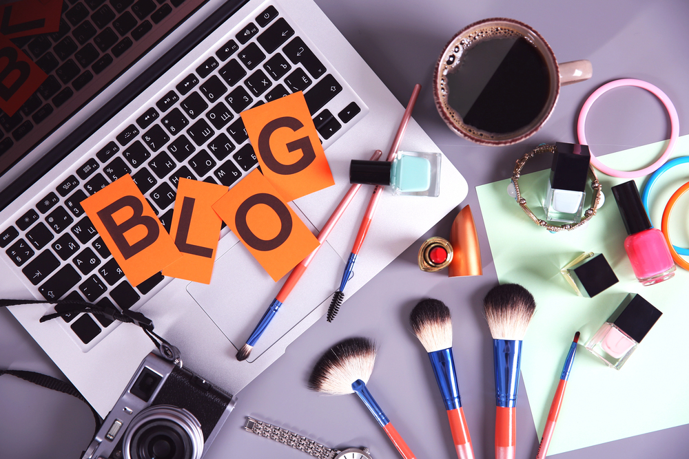 A laptop surrounded by make-up brushes, nail varnish, a camera, a cup of coffee, hair bands, and sticky notes spelling BLOG on the keyboard, suggesting beauty blogging or content creation.