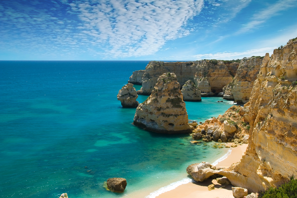 Clear turquoise water surrounds dramatic limestone cliffs and rock formations along a sunny coastline, with a sandy beach below and wispy clouds stretching across the bright blue sky.