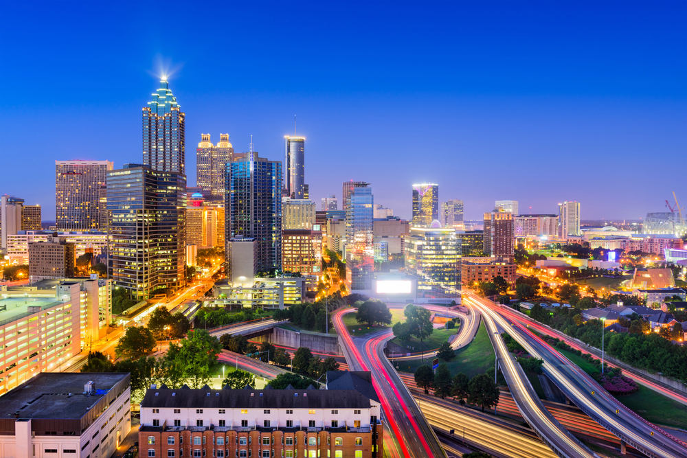 Atlanta city skyline at dusk with illuminated skyscrapers and bright lights. Blurred car lights show busy motorways in the foreground, while buildings and trees fill the urban landscape under a clear blue sky.