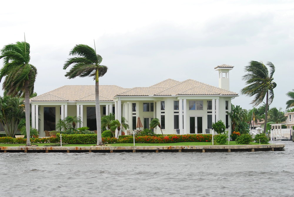 A large white waterfront house with tall windows and a tiled roof sits behind a small sea wall, surrounded by palm trees blown by strong wind, with choppy water in the foreground under a cloudy sky.