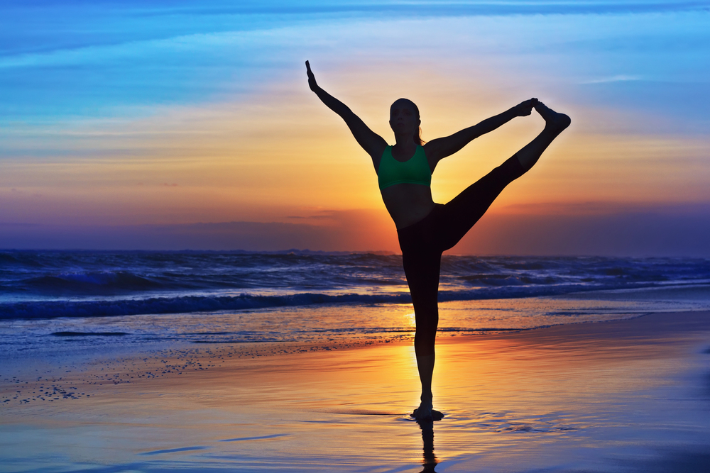 A person practises a yoga pose on a beach at sunset, standing on one leg with the other leg extended and one arm raised, silhouetted against the colourful sky and ocean.