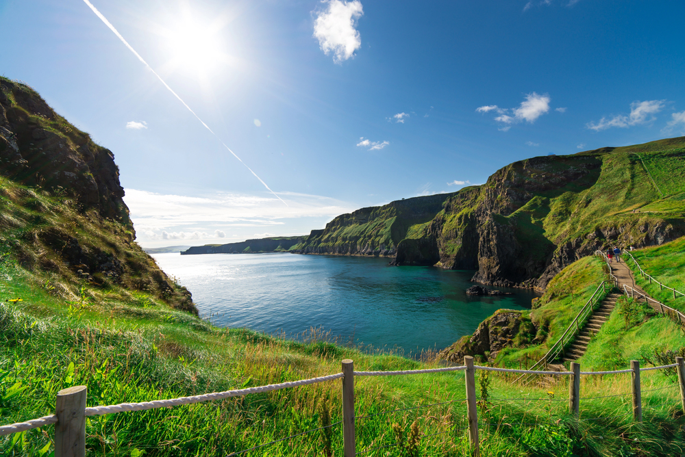 A sunny coastal landscape with green cliffs, blue water, clear sky, and a wooden path with rope railing winding down the hillside.