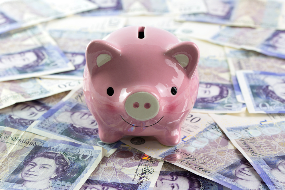A pink piggy bank sits on top of a scattered pile of British pound notes, symbolising savings and financial management.