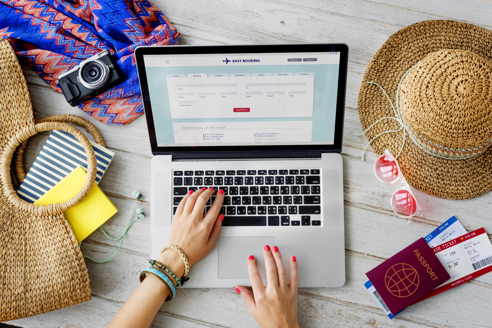A person using a laptop to book a holiday, surrounded by travel items including a straw hat, camera, sunglasses, woven bags, a passport, plane tickets, a notebook, and a colourful dress on a white wooden table.