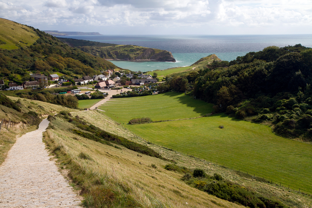 A paved path leads down a grassy hillside towards a small village and green fields, with cliffs and the sea in the background under a partly cloudy sky.