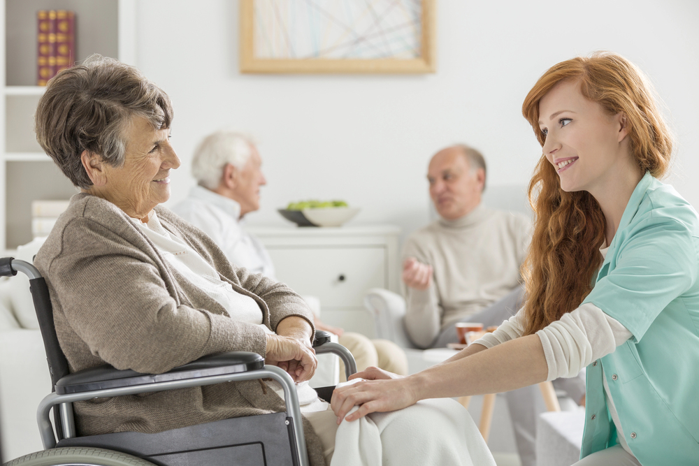 A young carer smiles whilst assisting an elderly woman in a wheelchair. In the background, two elderly men sit together and talk in a bright, comfortable room.