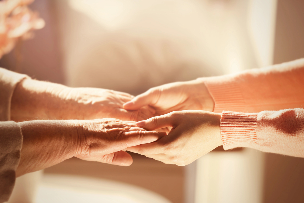 Close-up of a younger person gently holding the hands of an older person, conveying care, comfort, and support, with a warm, soft light in the background.