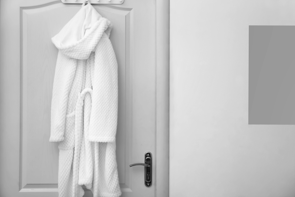A white bathrobe hangs on a hook over a closed white door with a silver handle; the door is set in a plain, light-coloured wall, with a rectangular grey patch visible on the right side of the image.