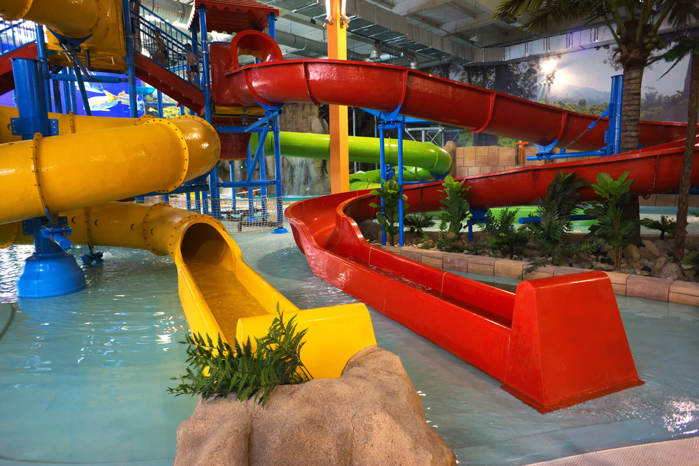 Indoor water park with winding yellow, red, and green water slides emptying into a shallow pool, surrounded by artificial plants, rocks, and tropical décor under a high ceiling.