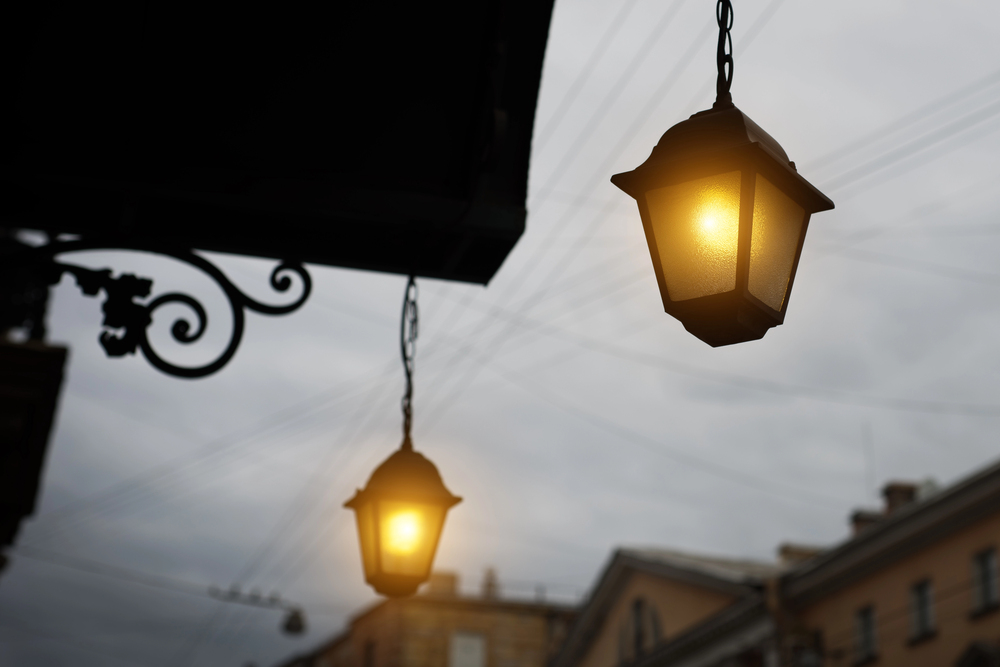 Two vintage-style street lamps glowing warmly at dusk, with a cloudy sky and blurred buildings in the background. Decorative metalwork is visible on the left side.