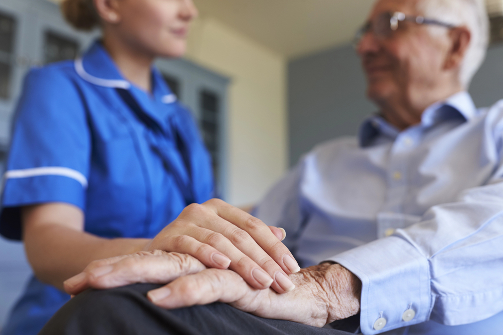 A nurse in blue uniform gently holds the hand of an elderly man wearing glasses and a light blue shirt, providing comfort and support in a calm indoor setting.