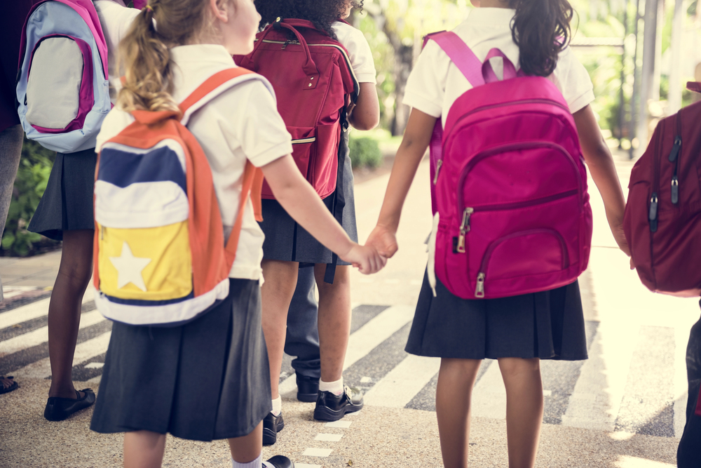 A group of school children in uniforms and rucksacks hold hands whilst walking across a zebra crossing outdoors.