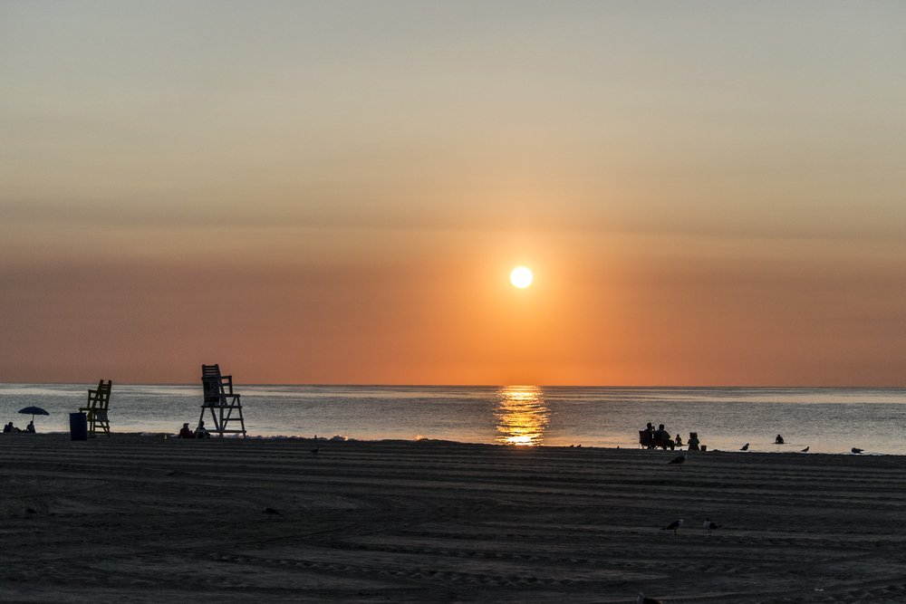 A peaceful beach at sunset with the sun low over the horizon, casting a golden reflection on the water. Silhouettes of people, lifeguard chairs, and birds are visible on the sand.