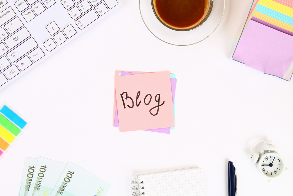 A pink sticky note with Blog written on it sits on a white desk surrounded by a keyboard, colourful sticky notes, a cup of coffee, euro notes, a notepad with a pen, and a small white alarm clock.