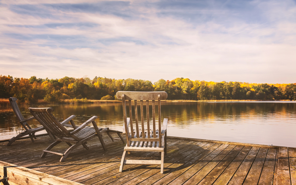 Wooden chairs sit on a jetty overlooking a calm lake, with trees in autumn colours reflecting in the water under a partly cloudy sky. The scene evokes a peaceful, relaxing atmosphere.