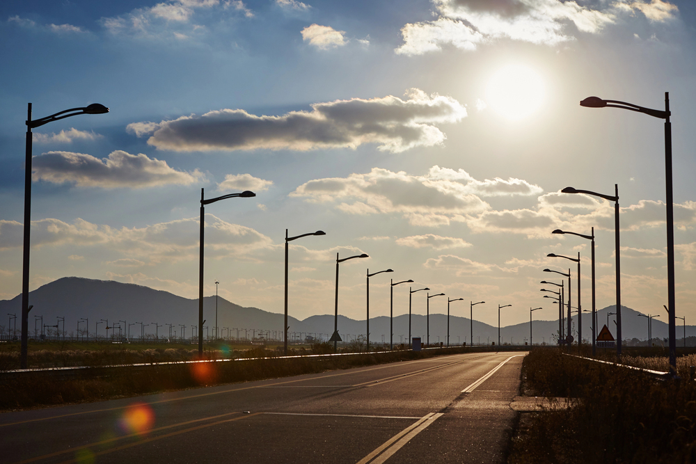 A wide, empty road lined with streetlights stretches into the distance under a bright sun and partly cloudy sky, with distant mountains silhouetted on the horizon.