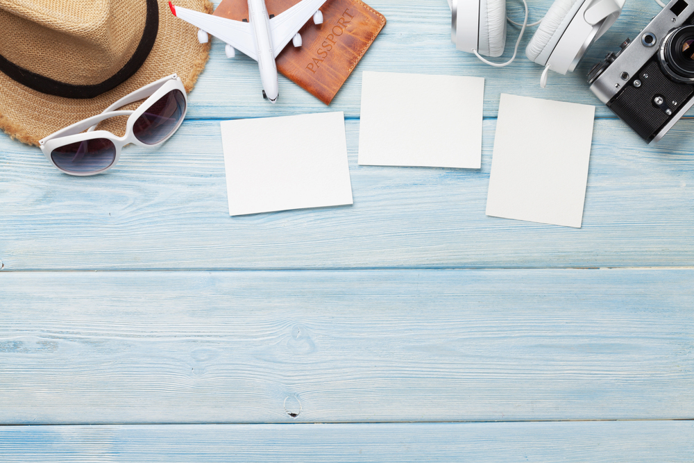 Travel-themed flat lay on blue wooden surface with a straw hat, sunglasses, toy aeroplane, passport, headphones, camera, and three blank white cards arranged in the centre.