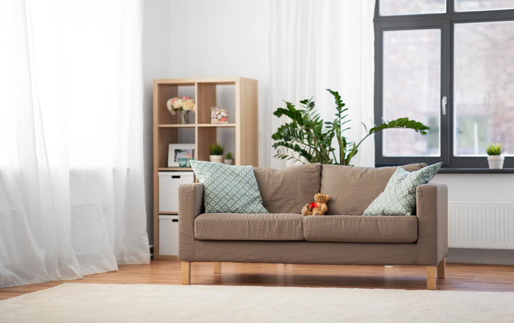 A beige sofa with two patterned blue cushions and a teddy bear sits in a bright lounge with a large window, light curtains, houseplants, and a wooden shelf with decorative items.