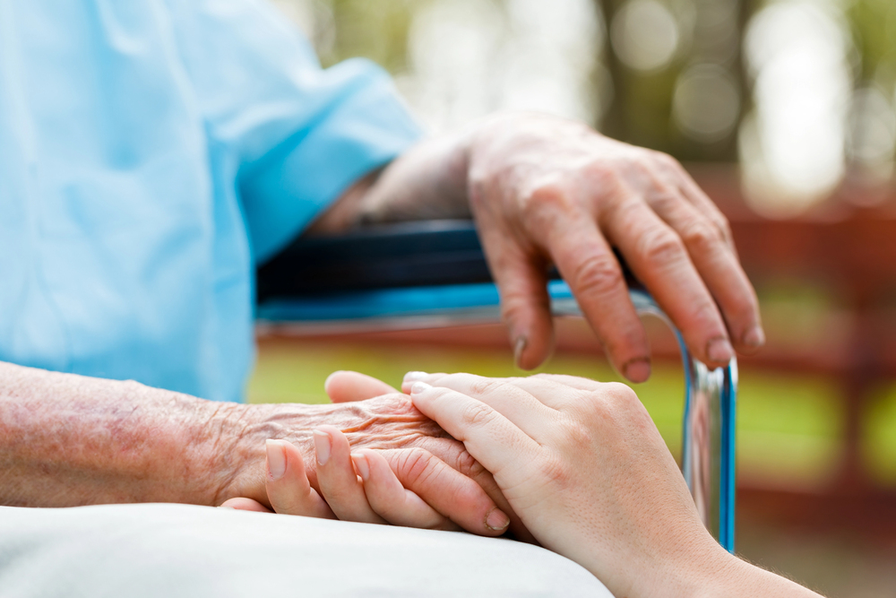 An elderly person in a wheelchair rests one hand on the armrest whilst another person gently holds their other hand, conveying comfort and support. Outdoor background is slightly blurred.