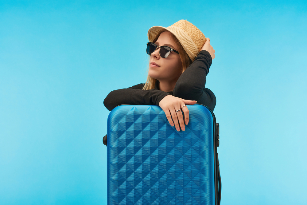 A woman wearing sunglasses and a straw hat leans on a blue textured suitcase against a bright blue background, looking upwards with a relaxed expression.
