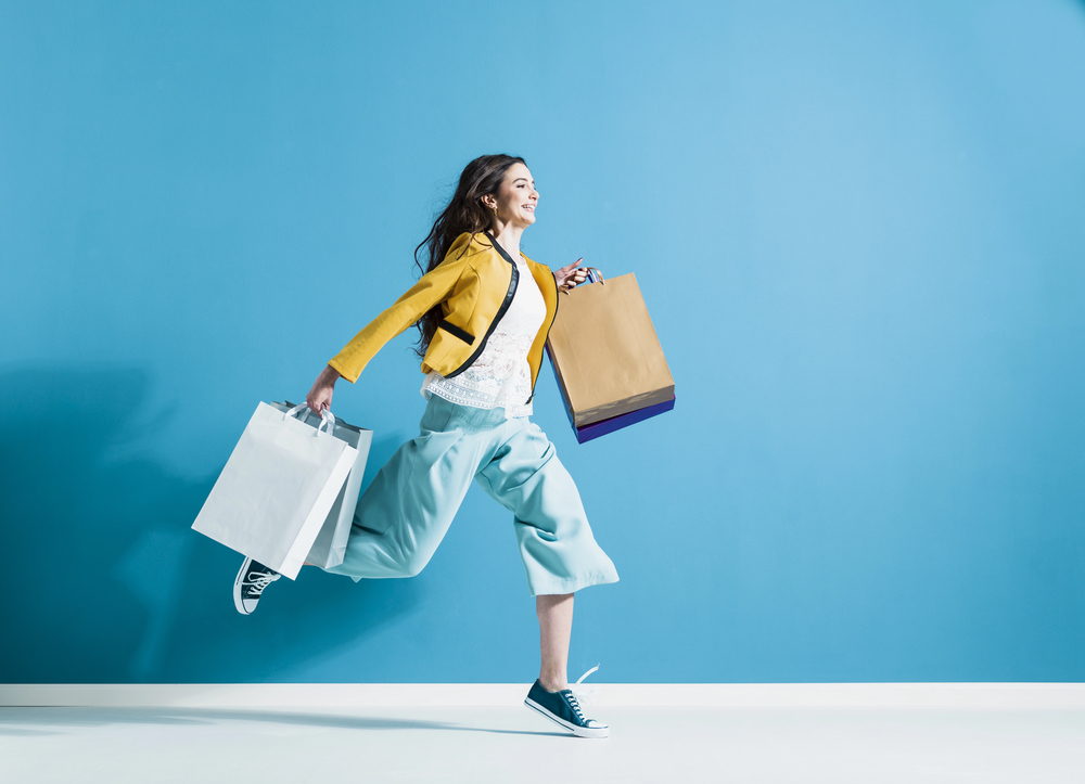 A woman in a yellow jacket and blue trousers happily runs with two shopping bags against a bright blue wall, smiling and wearing black trainers.