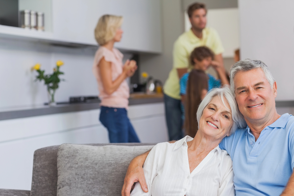 An older couple sits smiling on a sofa in a bright living room, while three adults and two children interact in the background near a kitchen worktop.