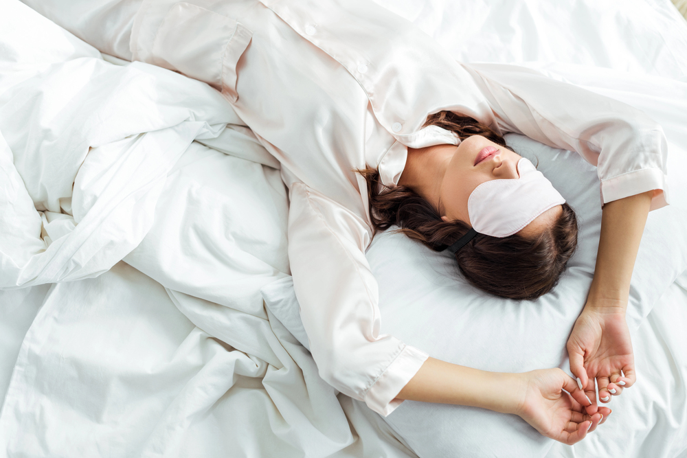 A woman wearing a silk sleep mask and matching pyjamas lies on her back in bed with white sheets, arms stretched above her head, appearing relaxed and comfortable.