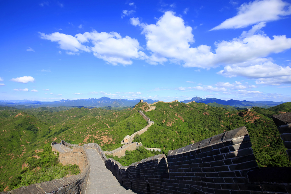 The Great Wall of China winds over lush green hills under a bright blue sky with scattered white clouds.