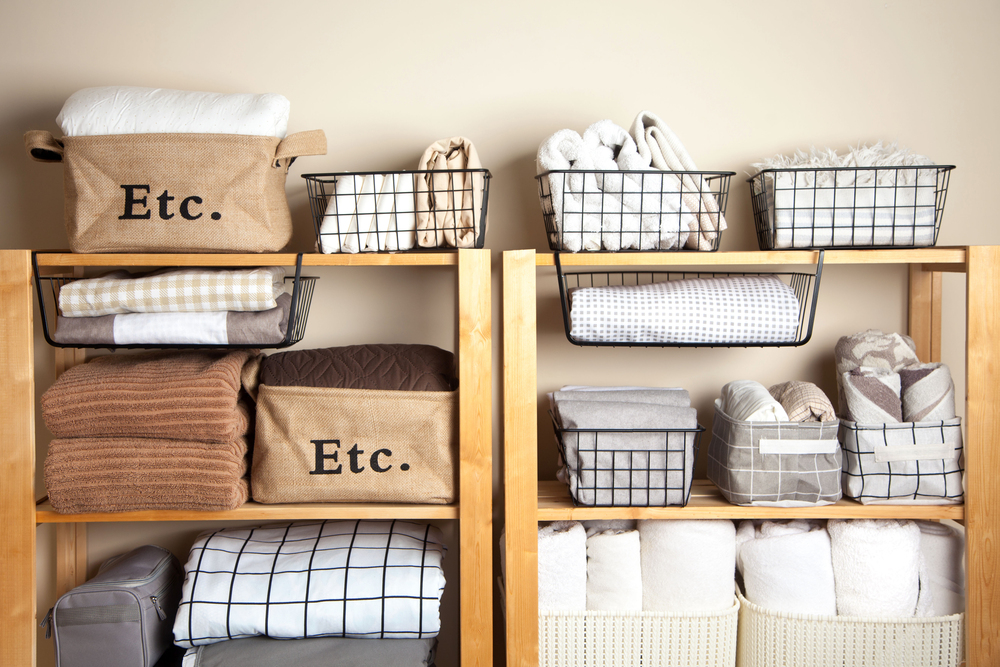Wooden shelves neatly organised with folded blankets, towels, and bins labelled Etc. and wire baskets holding rolled towels and other linens against a neutral wall.