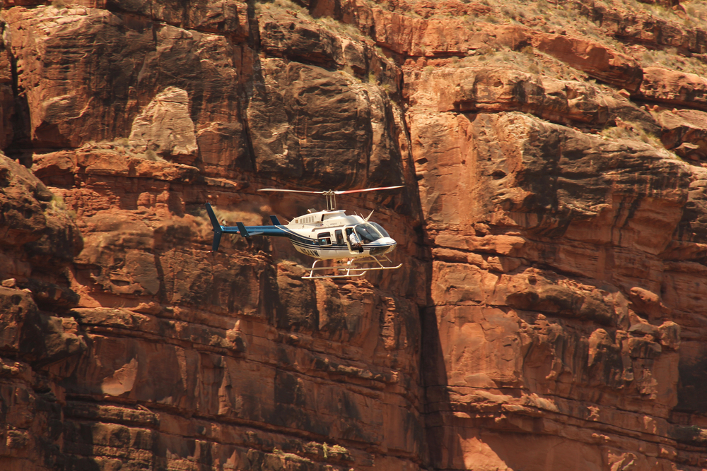 A white and blue helicopter flies in front of steep, rugged red rock cliffs, hovering close to the weathered canyon wall under bright daylight.
