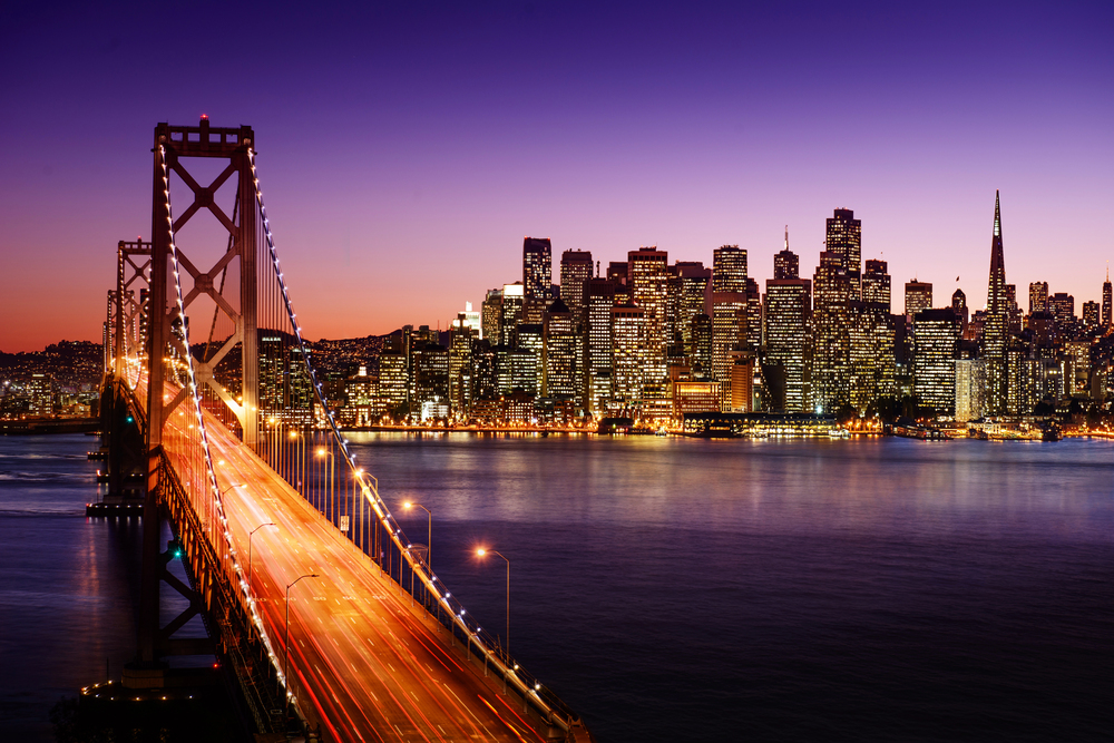 The San Francisco skyline at dusk, with the Bay Bridge in the foreground and city lights reflecting on the water under a purple and orange sky.