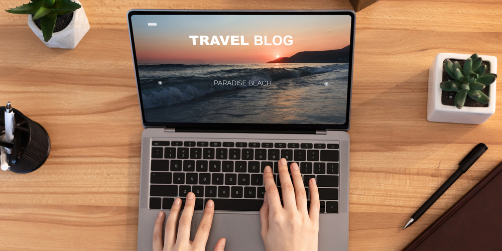 A person types on a laptop displaying a travel blog with a beach sunset photo on the screen. The desk also has a plant, a notebook, and a pen.