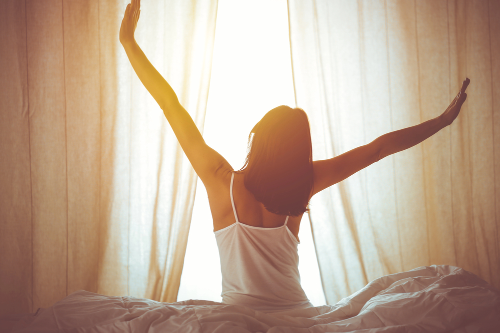 A woman sits on a bed, facing sunlit curtains, stretching her arms wide above her head. She wears a white vest, and the room glows softly with morning light.