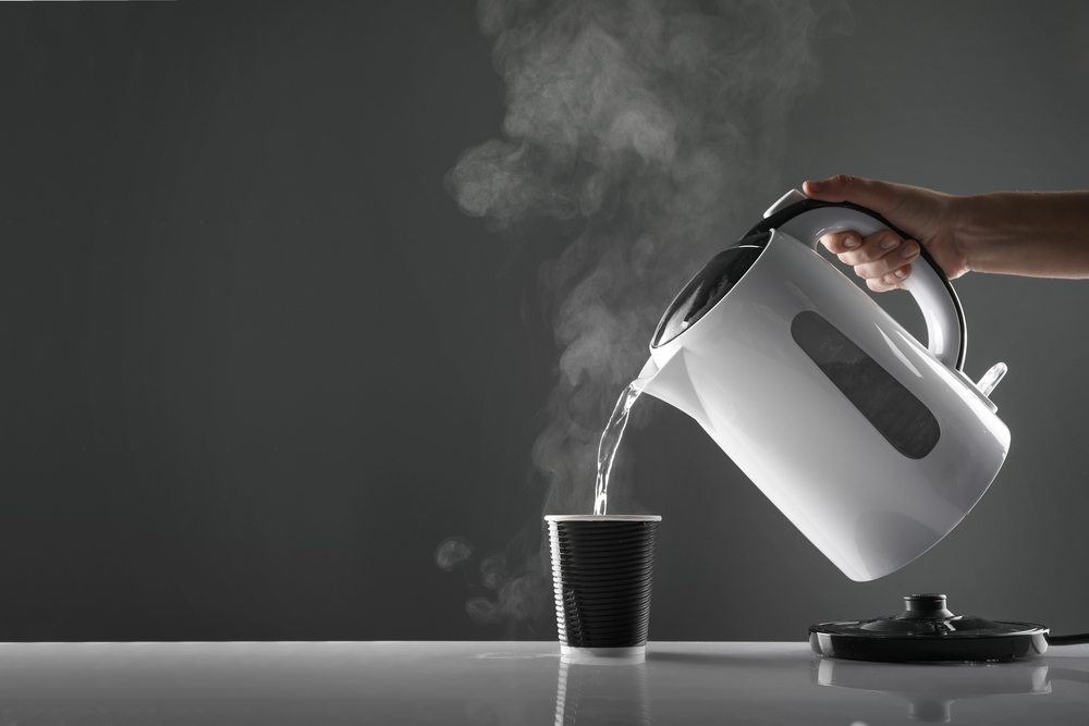 A hand pours steaming hot water from a white electric kettle into a black disposable cup on a glossy surface, with the kettle lid placed nearby against a dark grey background.