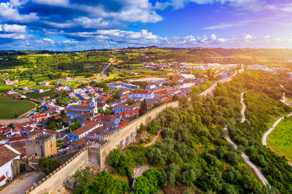 Aerial view of a historic walled town with red-roofed buildings, surrounded by lush green countryside and trees under a partly cloudy sky at sunset.