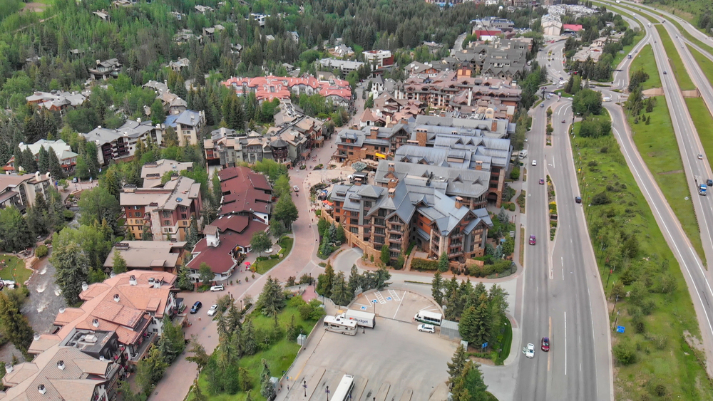 Aerial view of a small mountain town with clustered buildings, roads, and parked vehicles surrounded by green trees and nearby motorways.
