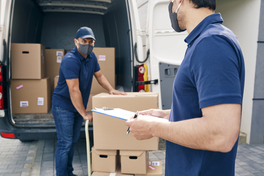 Two delivery workers wearing face masks unload cardboard boxes from a van. One worker pushes boxes on a trolley, while the other holds a clipboard and pen, checking items.