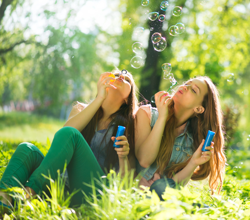 Two young women sit on grass in a sunny park, smiling and blowing soap bubbles. The background is filled with green trees and sunlight, creating a cheerful, relaxed atmosphere.
