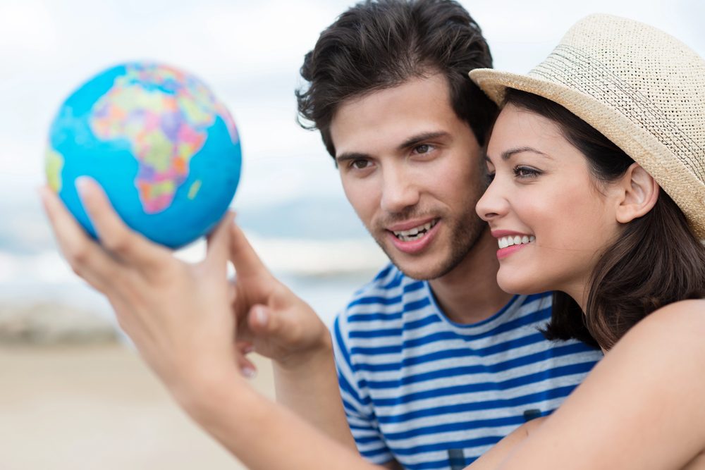 A smiling couple looks closely at a small globe, with the woman wearing a straw hat and the man in a striped shirt. The background is blurred, suggesting an outdoor or beach setting.