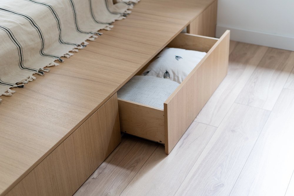 A wooden platform bed with built-in storage drawers, one drawer partially open revealing neatly folded bedding inside, sits on a light wood floor next to a bed with a striped beige blanket.