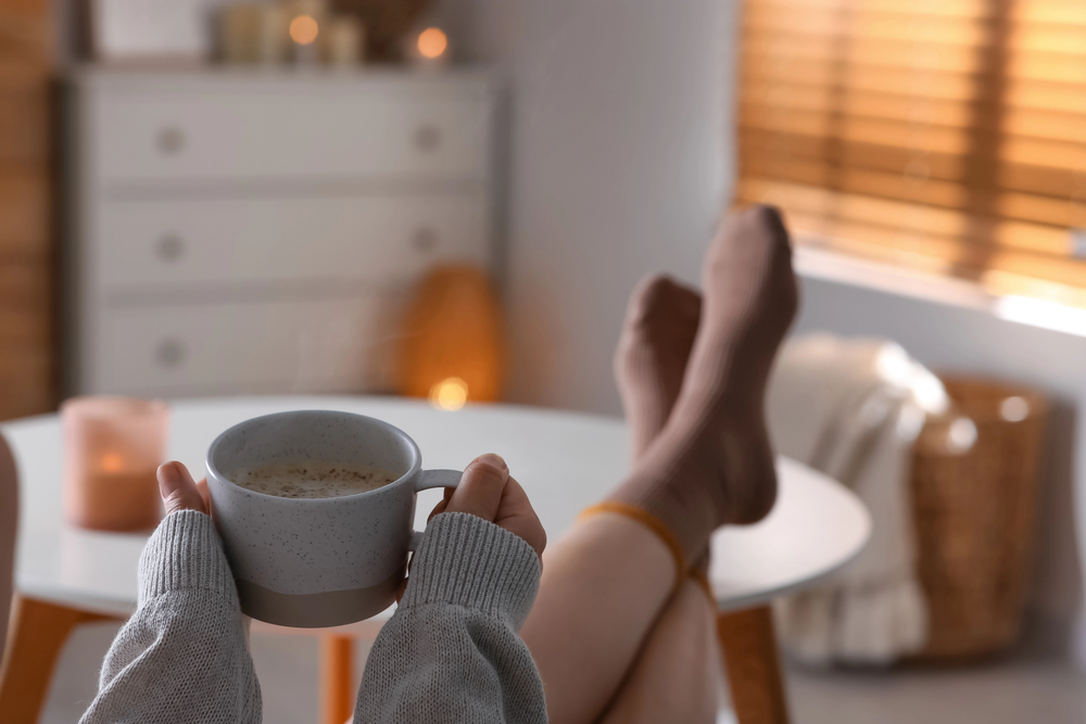A person wearing cosy socks relaxes with feet up on a table, holding a steaming mug, in a softly lit, warm room with candles, a basket, and wooden blinds in the background.