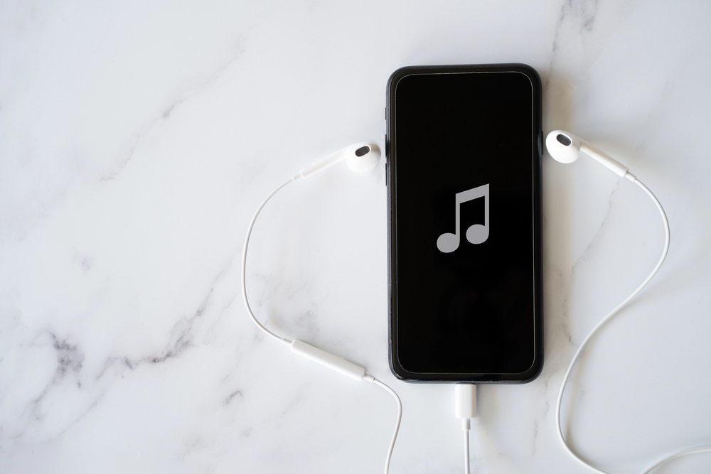 A smartphone with a music note icon on its screen is placed on a white marble surface, with white earphones plugged into it and lying beside the phone.