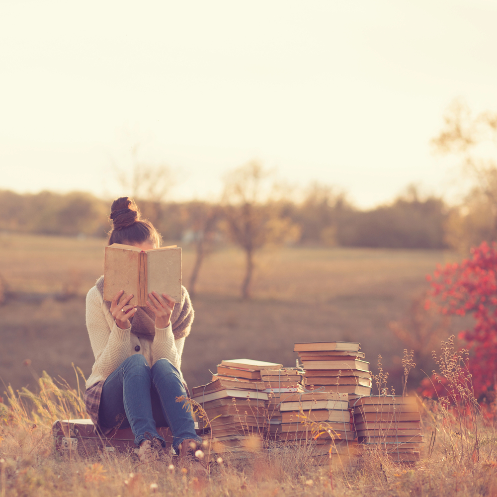A person sits outdoors on grass, reading a book with several stacks of books beside them. They wear a jumper and jeans, and the background features soft sunlight and autumn trees.